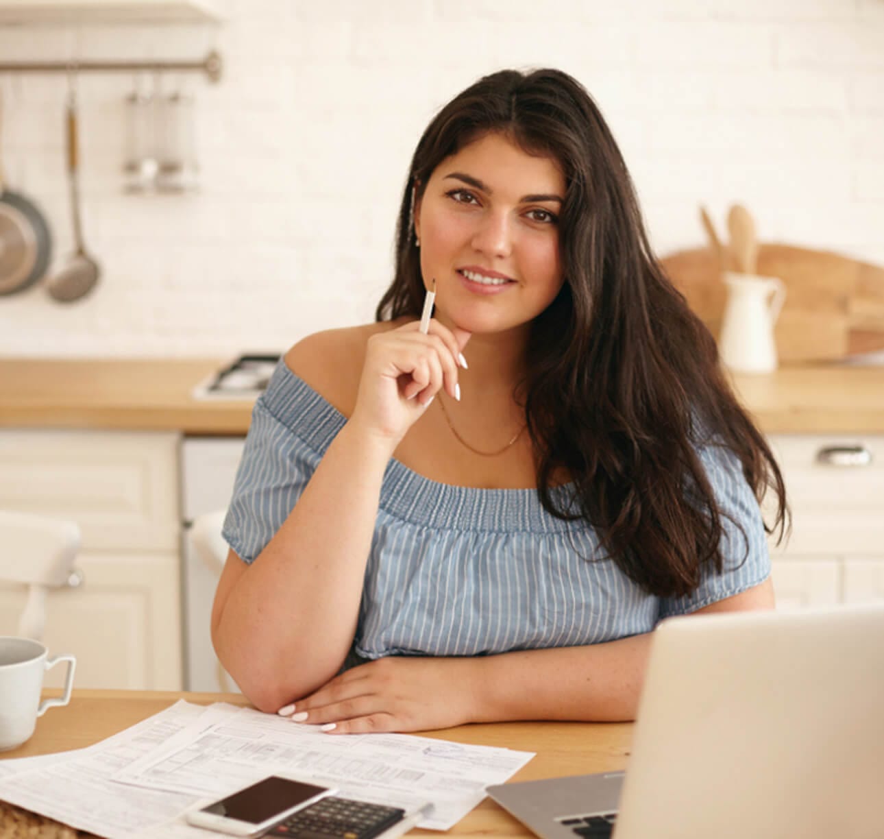 Smiling woman with long hair sitting at a kitchen table, working on finances with papers and a laptop.